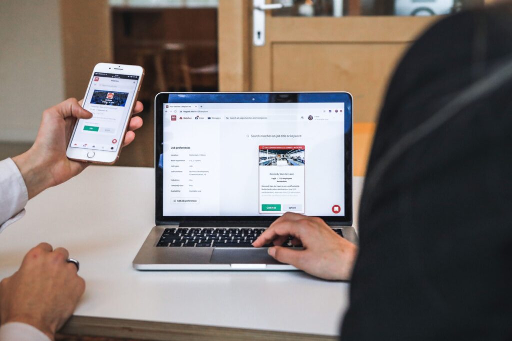 Two users comparing a website on a smartphone and laptop, illustrating how to confirm consistent domain, service, and account information before registering or paying.